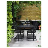 Modernes Esszimmer im Freien mit schwarzen Stühlen und Tisch auf einer Holzterrasse mit gelber Backsteinmauer im Hintergrund und üppigem Grün.