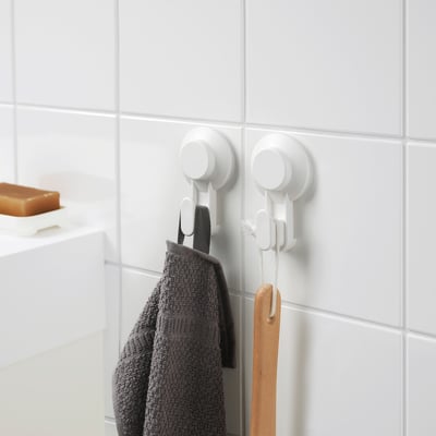 An einer gefliesten Wand hängen zwei weiße TISKEN-Haken, die ein graues Handtuch und ein Holzutensil halten, und zeigen so ihre Saugfähigkeit.