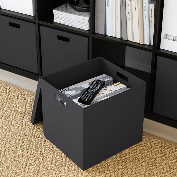 Schwarze TJENA-Box auf dem Boden, hält Fernbedienung und Box, passt auf das Kallax-Regal.