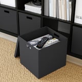 Schwarze TJENA-Box auf dem Boden, hält Fernbedienung und Box, passt auf das Kallax-Regal.