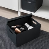 Schwarze TJENA-Aufbewahrungsbox mit Kopfhörern und einem Konsolen-Controller, gezeigt auf einem Teppichboden in der Nähe eines weißen Schreibtisches.