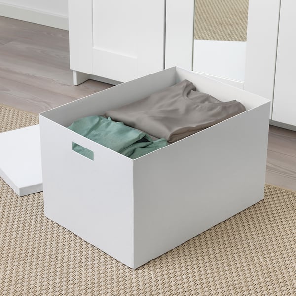 Eine weiße TJENA-Aufbewahrungsbox mit ausgeschnittenen Griffen, gefüllt mit gefalteten grauen und grünen Kleidungsstücken, die auf einem gewebten Teppich platziert sind.