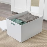 Eine weiße TJENA-Aufbewahrungsbox mit ausgeschnittenen Griffen, gefüllt mit gefalteten grauen und grünen Kleidungsstücken, die auf einem gewebten Teppich platziert sind.