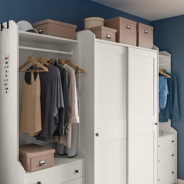 Ein gut organisierter Kleiderschrank mit weißen TJOG-Boxen, in denen Kleidung und Gegenstände auf Schränken verstaut sind, vor einer dunkelblauen Wand.
