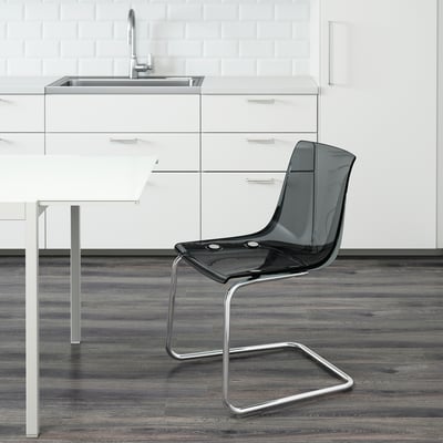 Moderne Küche mit schlanken Schränken und Spüle. Im Vordergrund transparenter TOBIAS-Stuhl mit flexibler Sitzfläche und Rückenlehne, der sich in die Umgebung einfügt.