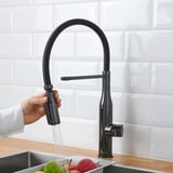 Eine Hand hält eine schwarze Küchenarmatur mit Handspray und spült Gemüse unter sauberem Wasser ab. Die Armatur ist ein ausziehbarer Sprüher für die praktische Reinigung.