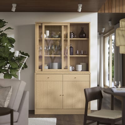 Holzschrank mit Glastüren, Regalen und Soft-Close-Schubladen. Umgeben von Stühlen, in der Nähe einer großen Pflanze.