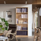 Bücherregal aus Eichenholz mit Büchern und Dekor, neben dunklem Stuhl, Pflanze und moderner Malerei.