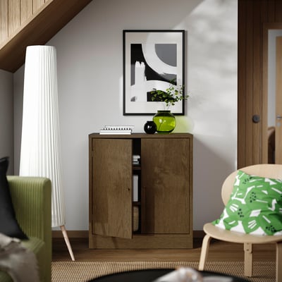 Brauner Schrank, grüne Vase mit Blumen, Bücher. Graue Wand, Gemälde. Lampe links, gemusterter Stuhl rechts. Gemütliches Zimmer.