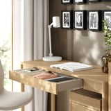 Schreibtisch aus Holz mit offener Schublade für Aktenordner und Notizbücher. Oben befinden sich eine Lampe, eine Pflanze und ein Fotorahmen.
