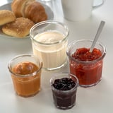Kleine Gläser gefüllt mit verschiedenen Marmeladen und einer Tasse Sahne neben Brötchen, ideal für Frühstücksaufstriche.