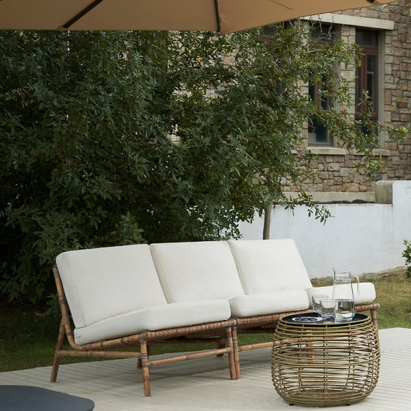 Weißes TVARÖ-Sofa für den Außenbereich, groß, mit beigen Kissen, auf der Terrasse in der Nähe des Hauses, neben kleinem Tisch mit Getränken.