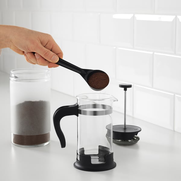Eine Person gibt Kaffeesatz in eine durchsichtige UPPHETTA French Press. Daneben ist der Kolben zu sehen, der auf einer Theke verwendet werden kann.