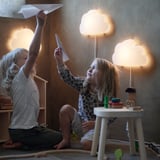 Kinder spielen mit Papierfliegern unter Wolkenlampen. Einer in Weiß, der andere in Leopardenmuster, Spielzeug in der Nähe.