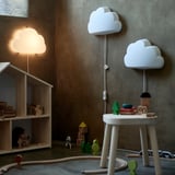 Verspieltes Kinderzimmer mit weißen, wolkenförmigen Lampen, einer leuchtenden, weichen Lichtlampe, Holzspielzeug und einem Puppenhaus, das eine gemütliche und fantasievolle Atmosphäre schafft.