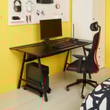 UTESPELARE Gaming-Schreibtisch in Schwarz, mittige Mittelposition, mit Monitor & Zubehör, rot-schwarzer ergonomischer Stuhl, gelber Wandhintergrund.