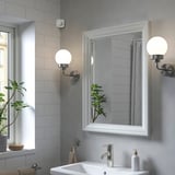 Modernes Badezimmer mit VALLHORN-Bewegungsmelder über weißem Waschbecken, silbernem Wasserhahn, Spiegel und Pflanze. Hellgrau geflieste Wände.