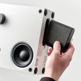 Eine Person verbindet ein schwarzes Gerät mit der Rückseite eines weißen VAPPEBY-Bluetooth-Lautsprechers.