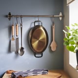Küchenszene mit Bräter aus Kohlenstoffstahl an Haken, Holz- und Metallbesteck und kariertem Stoff auf Holz.