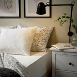 Schlafzimmerecke mit VÅRVIAL Bettwäsche in Creme. Schwarze verstellbare Lampe hängt über dem Nachttisch mit Buch und Vase. Kunstrahmen schmücken die Wand.