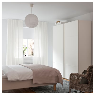 Schlafzimmer mit weißen Wänden, einem Bett mit brauner und weißer Bettwäsche, einem Korbstuhl und einer Hängeleuchte. Großes Fenster mit durchsichtigen Vorhängen.