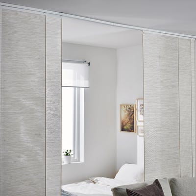 Schlafzimmer mit großen VIDGA-Schiebepaneelen in heller Farbe, die Fenster und Schrank abdecken. Die Paneele erscheinen höher als das Bett, das teilweise sichtbar ist.