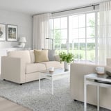 Minimalistisches Wohnzimmer: beiges VIMLE-Sofa, weißer Couchtisch, großes Fenster, helle Vorhänge, Blick auf die Natur.