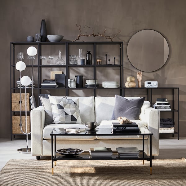 Modernes Wohnzimmer mit VIMLE-Schlafsofa, geometrischen Kissen, Glastisch, Regalen, Spiegel und strukturierter Wand.