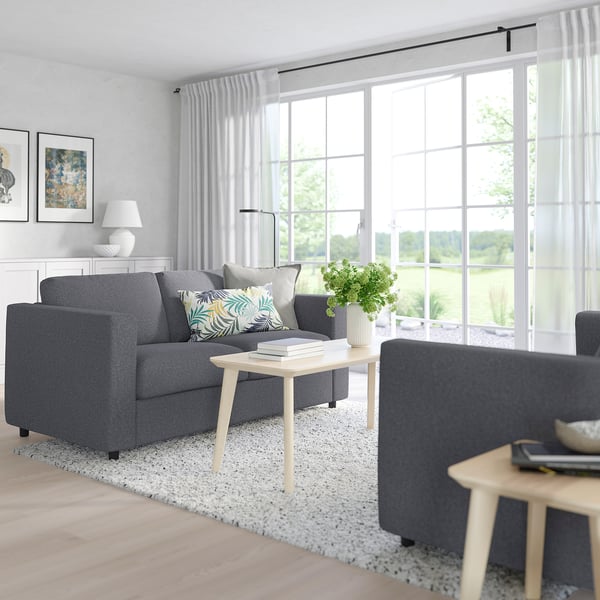 Modernes Wohnzimmer mit grauem VIMLE-Sofa, Couchtisch aus hellem Holz und großen Fenstern. Das Sofa ist groß und bietet Platz für mehrere Kissen mit ausreichend Sitzgelegenheiten.