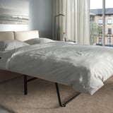 Ein modernes Schlafzimmer mit einem grauen VIMLE-Schlafsofa mit einer dicken Matratze vor einem großen Fenster mit Blick auf die Stadt, minimalistische Einrichtung.