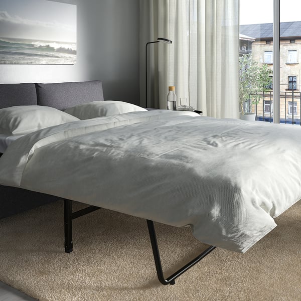 Bett mit weißem Bezug, grauen Kissen, schwarzen Metallbeinen. Großes Fenster, Blick auf die Stadt, Vorhang, Nachttisch mit Lampe und Gemälde.