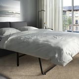 Modernes Schlafzimmer mit VIMLE-Schlafsofa, großem Fenster, weicher weißer Bettwäsche, dunklen Kissen, Stehlampe und Stadtblick.