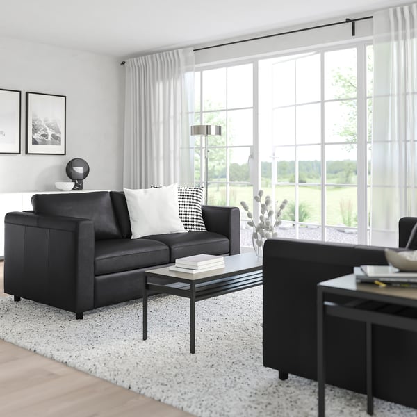 Modernes Wohnzimmer mit grauem VIMLE-Sofa, weißem Teppich, schwarzem Couchtisch und großem Fenster mit durchsichtigen Vorhängen.
