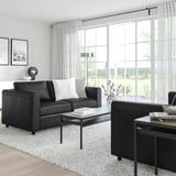 Modernes Wohnzimmer mit grauem VIMLE-Sofa, weißem Teppich, schwarzem Couchtisch und großem Fenster mit durchsichtigen Vorhängen.