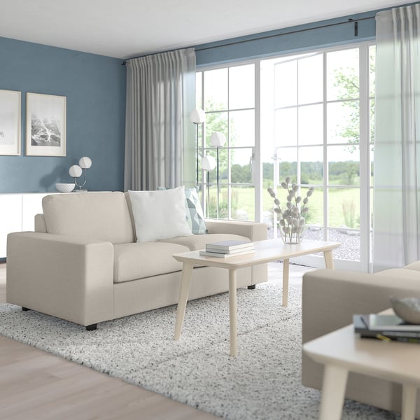 Modernes Wohnzimmer mit beigem VIMLE-Sofa, blauer Wand, weißen Kissen, Couchtisch aus Holz, Vase, Büchern und großen Fenstern.