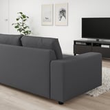 Modernes Wohnzimmer mit schwarzem VIMLE-Sofa, Flachbild-TV, Kunstwerken, Pflanzen und gesprenkeltem Teppich. Das Sofa kontrastiert mit hellen Wänden und Holzboden.