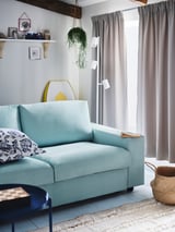 Ein gemütliches Wohnzimmer mit einem hellblauen VIMLE-Sofa, einem gemusterten Kissen, einer Hängepflanze und grauen Vorhängen.