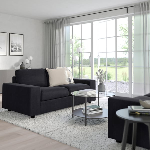 Modernes Wohnzimmer mit dunkelgrauem VIMLE-Sofa, Couchtischen aus Glas, Teppich, Lampe, Kunst und Fenster.