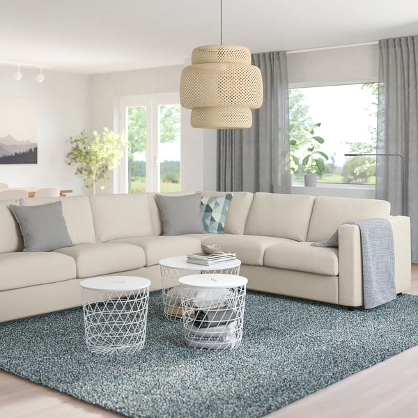 Modernes Wohnzimmer mit cremefarbenem VIMLE-Sofa, grauen Kissen, drei weißen Couchtischen auf blauem Teppich, abgestuftem Licht, großen Fenstern, Pflanzen.