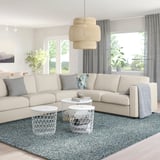 Modernes Wohnzimmer mit cremefarbenem VIMLE-Sofa, grauen Kissen, drei weißen Couchtischen auf blauem Teppich, abgestuftem Licht, großen Fenstern, Pflanzen.