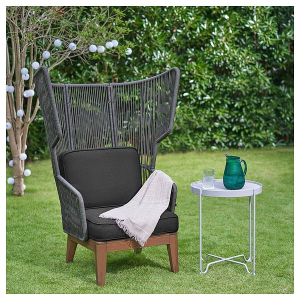 Ein moderner Outdoor-Stuhl, VINGSÖN, hohe Rückenlehne, dunkle Kissen, Holzbeine und flexibles Seilgeflecht. Daneben ein kleiner Beistelltisch mit grünem Krug & Glas.