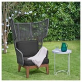 Ein moderner Outdoor-Stuhl, VINGSÖN, hohe Rückenlehne, dunkle Kissen, Holzbeine und flexibles Seilgeflecht. Daneben ein kleiner Beistelltisch mit grünem Krug & Glas.