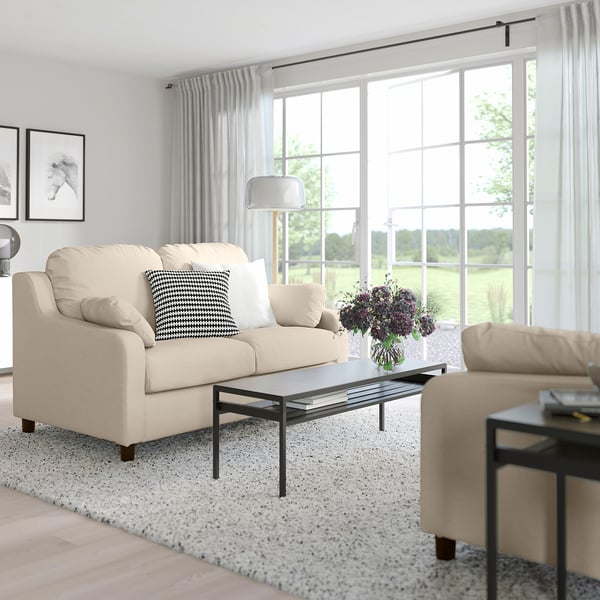Modernes Wohnzimmer mit beigem VINLIDEN-Sofa, schwarzem Couchtisch, Beistelltisch, Teppich und Blumenvase. Große Fenster mit durchsichtigen Vorhängen.
