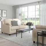 Modernes Wohnzimmer mit beigem VINLIDEN-Sofa, schwarzem Couchtisch, Beistelltisch, Teppich und Blumenvase. Große Fenster mit durchsichtigen Vorhängen.