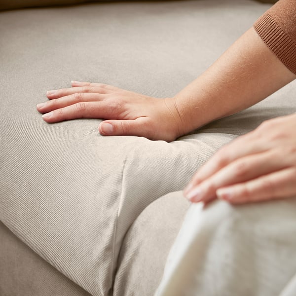 Person sitzt auf beigem VINLIDEN-Sofa, die Hände auf den Knien. Das Sofa hat weiche Kissen und einen glatten Stoffbezug.
