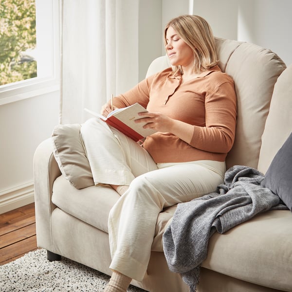 Frau liest auf beigem VINLIDEN-Sofa in der Nähe des Fensters. Sie trägt einen pfirsichfarbenen Pullover und eine leichte Hose. Eine graue Decke drapiert den Arm.