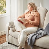 Frau liest auf beigem VINLIDEN-Sofa in der Nähe des Fensters. Sie trägt einen pfirsichfarbenen Pullover und eine leichte Hose. Eine graue Decke drapiert den Arm.