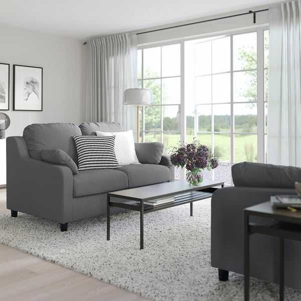 Modernes Wohnzimmer mit grauem VINLIDEN-Sofa, Couchtisch aus Glas, Beistelltisch und großem Fenster mit Vorhängen.