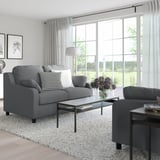 Modernes Wohnzimmer mit grauem VINLIDEN-Sofa, Couchtisch aus Glas, Beistelltisch und großem Fenster mit Vorhängen.