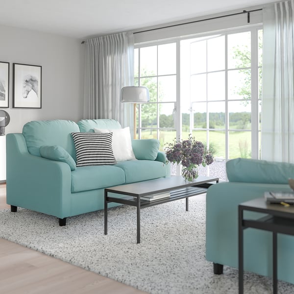 Wohnzimmer mit blauem Sofa und Sessel, Kissen, Holzboden, Fenster, Vorhängen, Couchtisch, Lampe, Beistelltisch, Teppich.